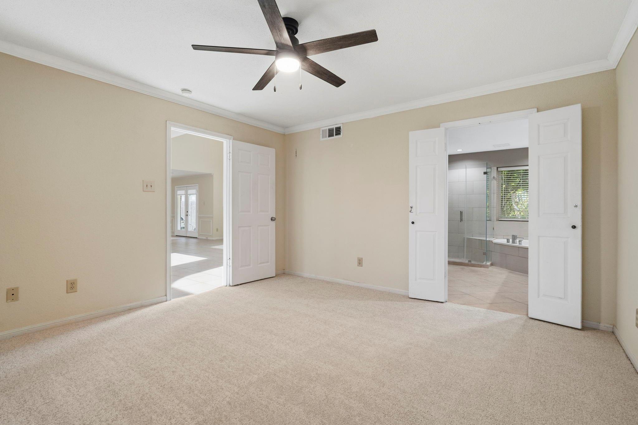 Empty carpeted bedroom with a ceiling fan, open to an ensuite bathroom and a bright tiled room.