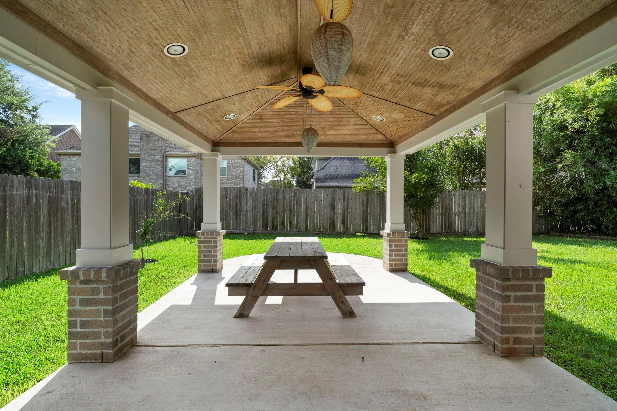 Covered backyard patio with a vaulted wood ceiling, ceiling fan, and picnic table.
