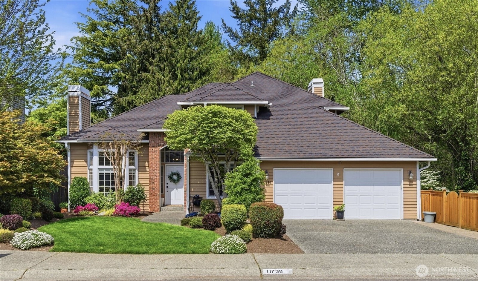 Tan house with brick entry, two-car garage, landscaped lawn, and mature trees.