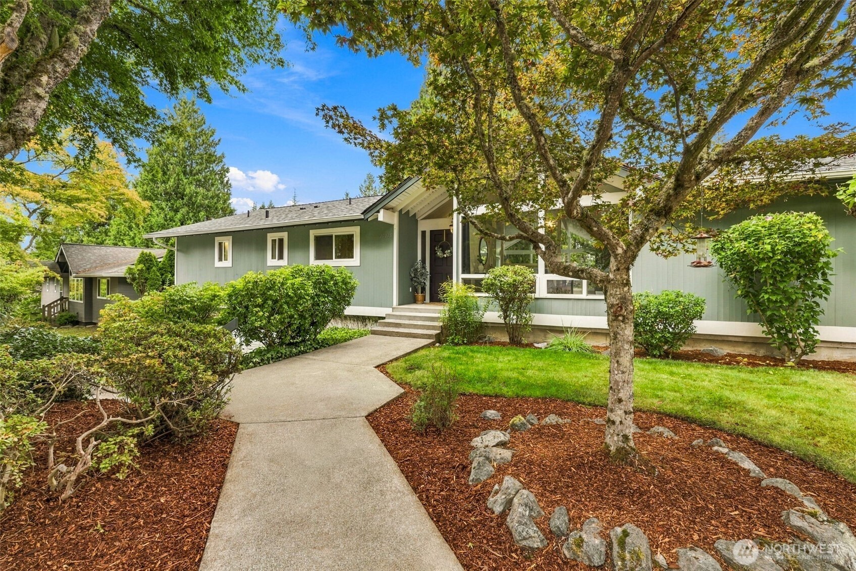 Green single-story home with a curved walkway, landscaped garden, and mature shade trees.