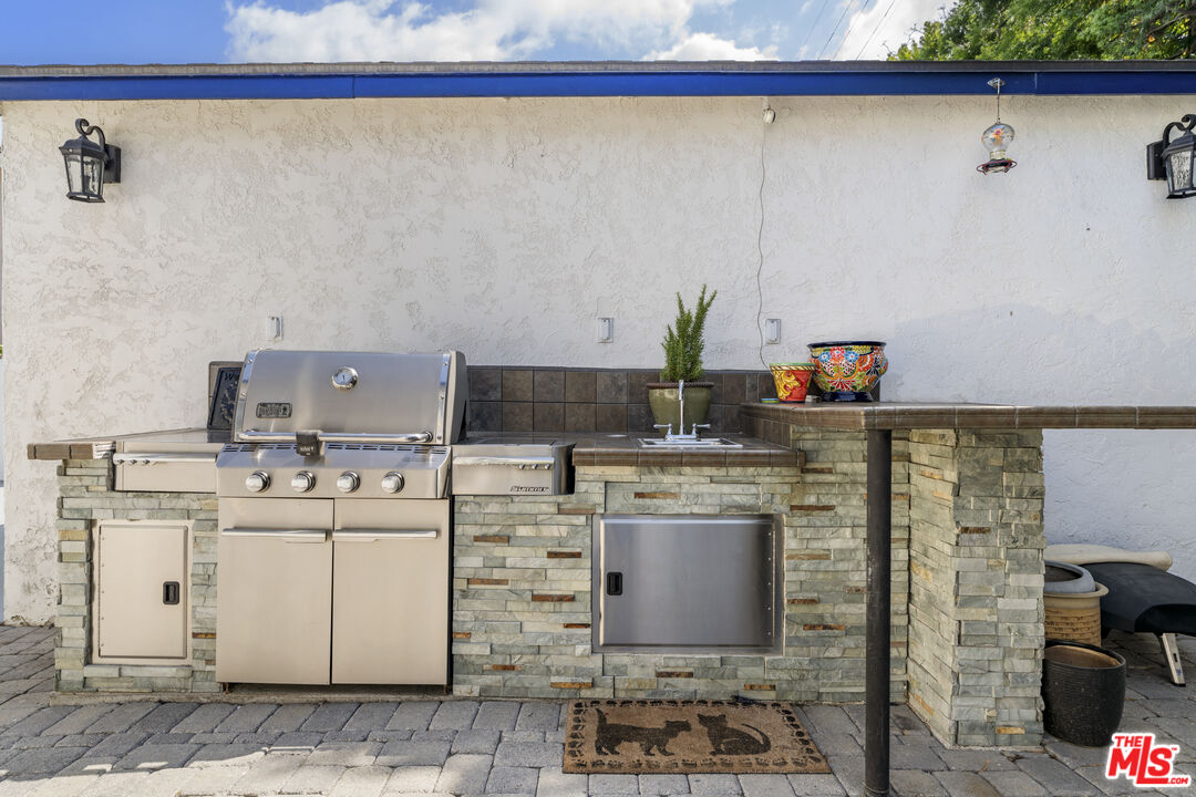Outdoor kitchen with a built-in stainless-steel grill, stone counters, sink, and raised bar.