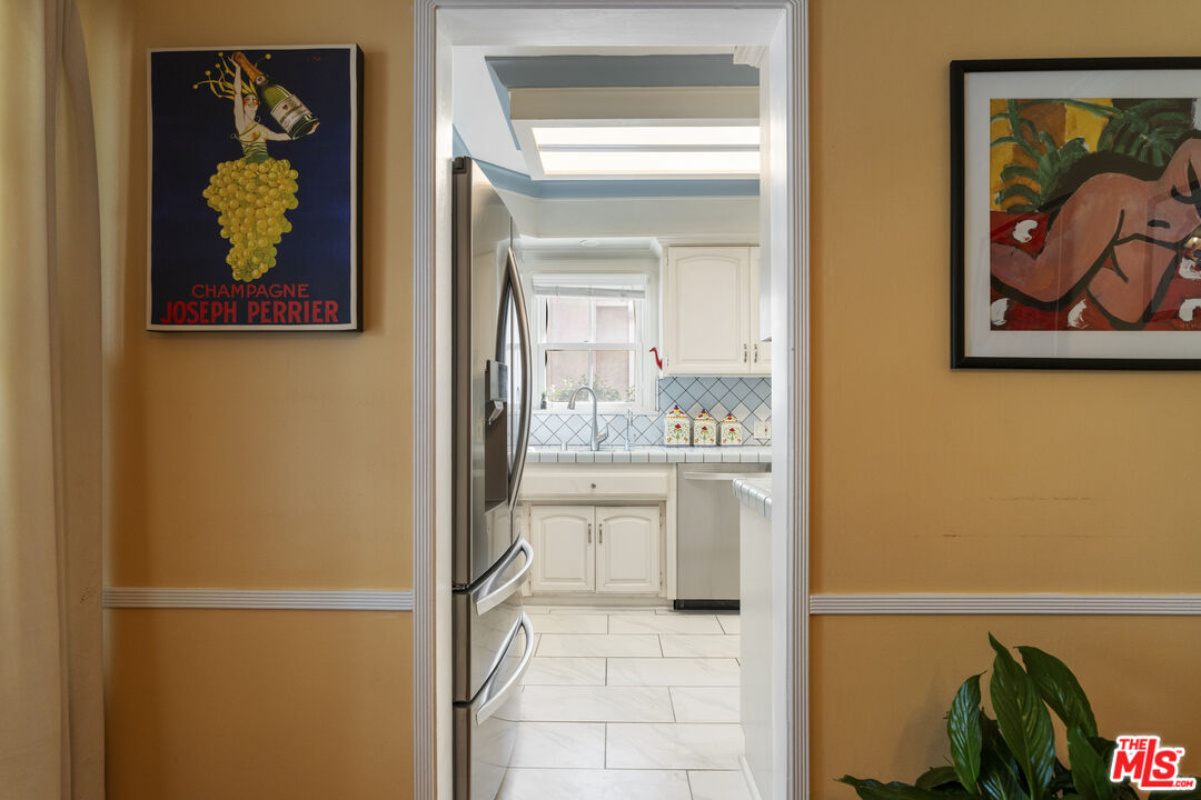 Bright white kitchen viewed through a doorway, with a stainless-steel refrigerator and diamond-pattern backsplash.