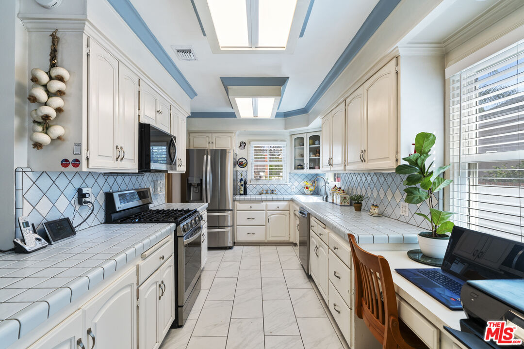 Bright galley kitchen with white cabinetry, tiled counters, stainless steel appliances, and a desk beside the window.