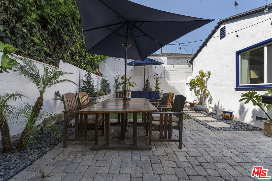 Outdoor dining patio with a wooden table, dark umbrellas, string lights, and potted plants.