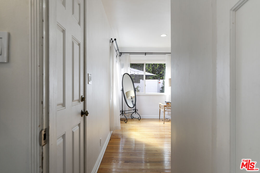 White hallway opens into a bright room with hardwood floors, a large window, and an oval mirror.