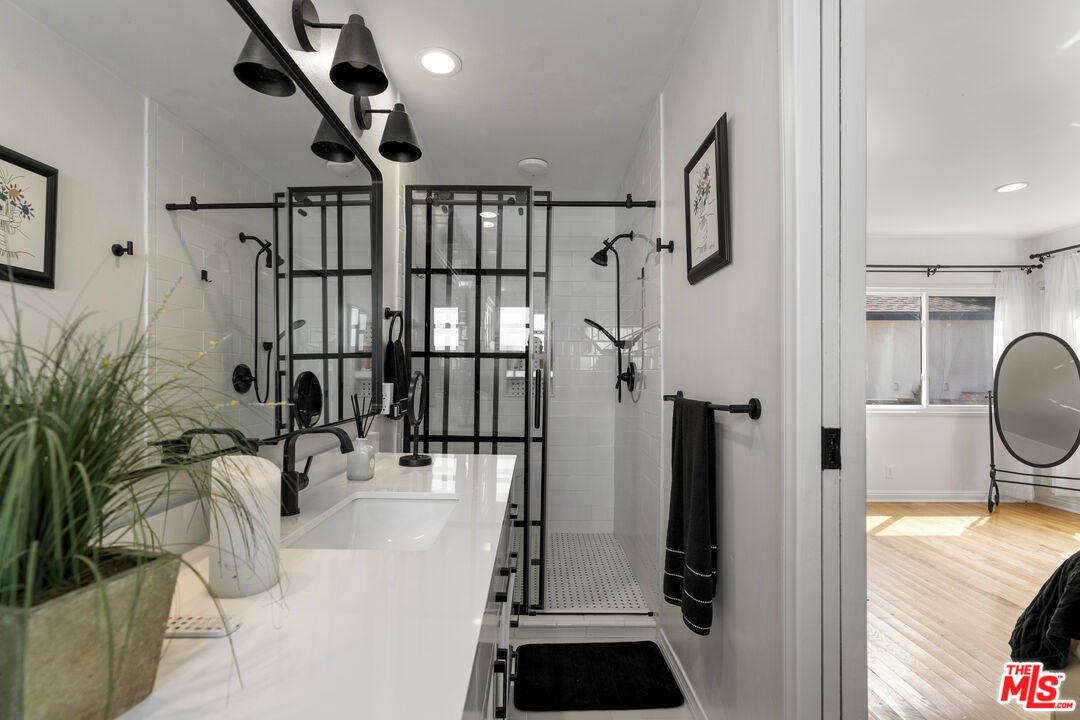 Bathroom with a black-framed glass shower, white vanity, and view into an adjoining hardwood-floored room.
