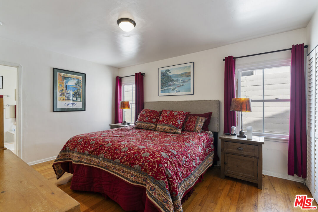 Bedroom with red patterned bedding, a gray upholstered headboard, hardwood floors, and burgundy curtains.