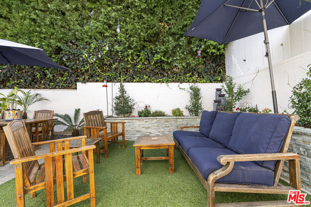 Outdoor patio with navy-cushioned sofa, wooden dining set, umbrellas, and a leafy privacy wall.