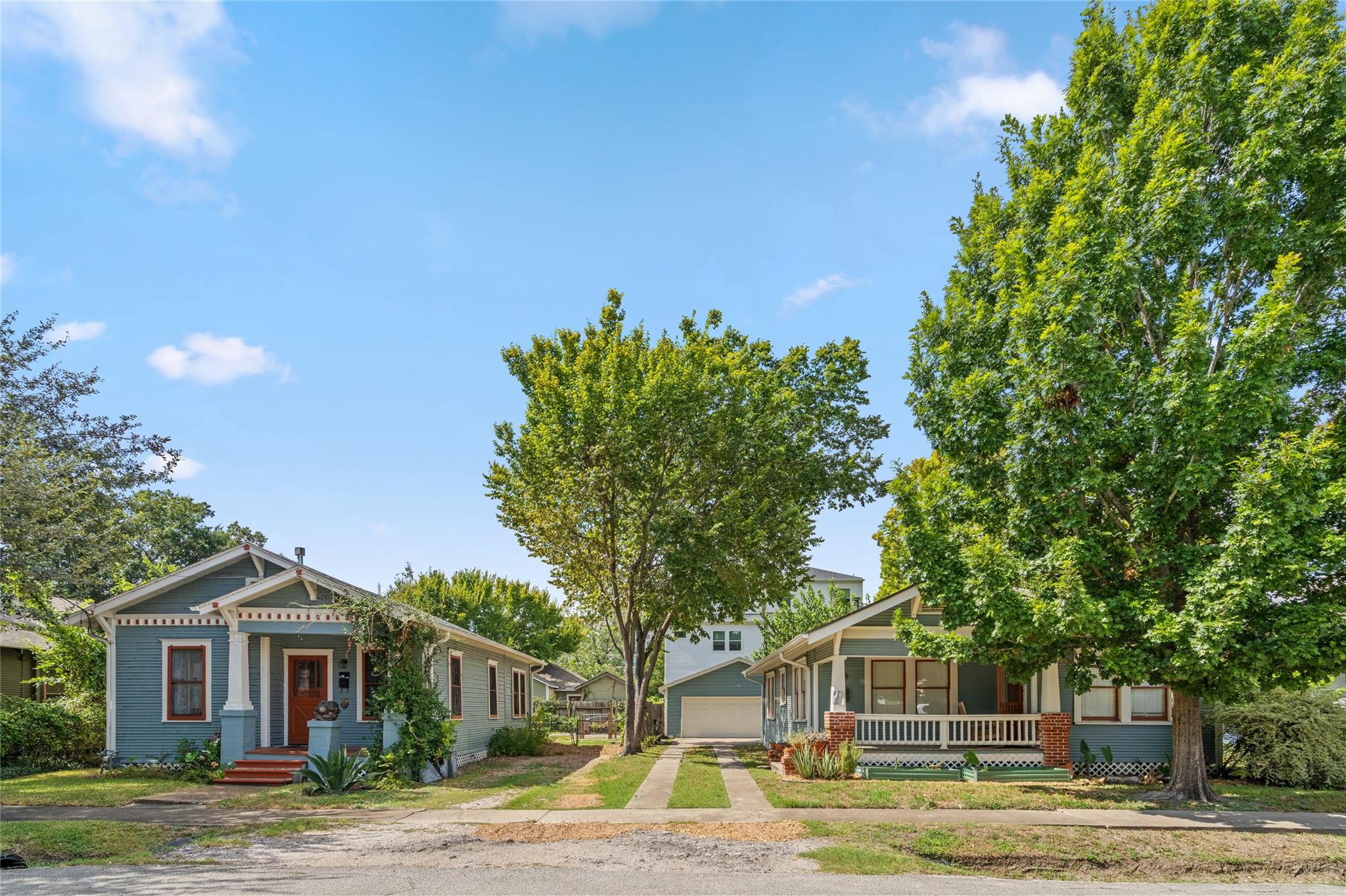 Two blue-gray Craftsman homes with porches stand beneath mature trees along a sunny residential street.