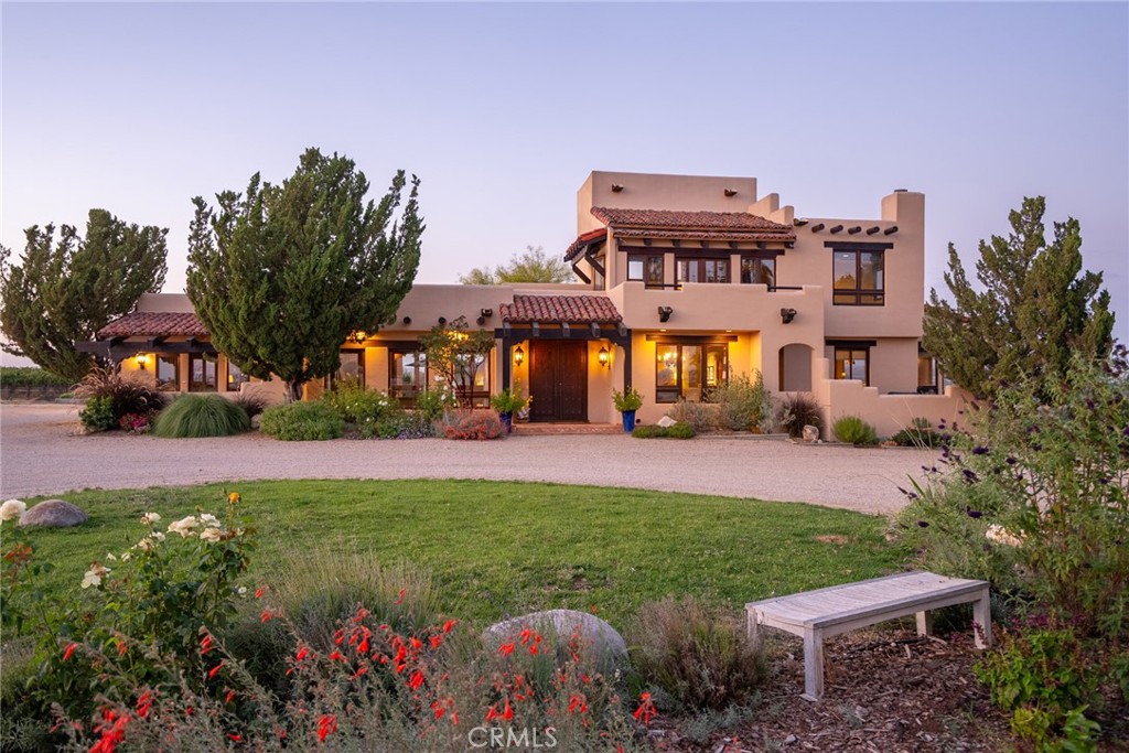 Two-story stucco home with an illuminated entry, landscaped garden, gravel drive, and wooden bench at dusk.