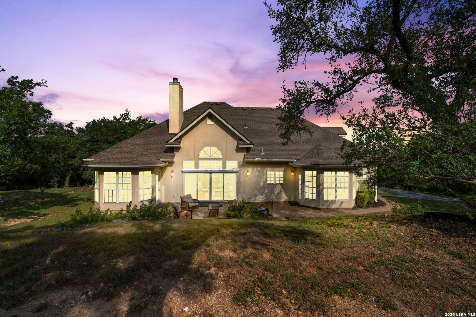 Rear of a beige house with broad lit windows and patio seating beneath a pink dusk sky.
