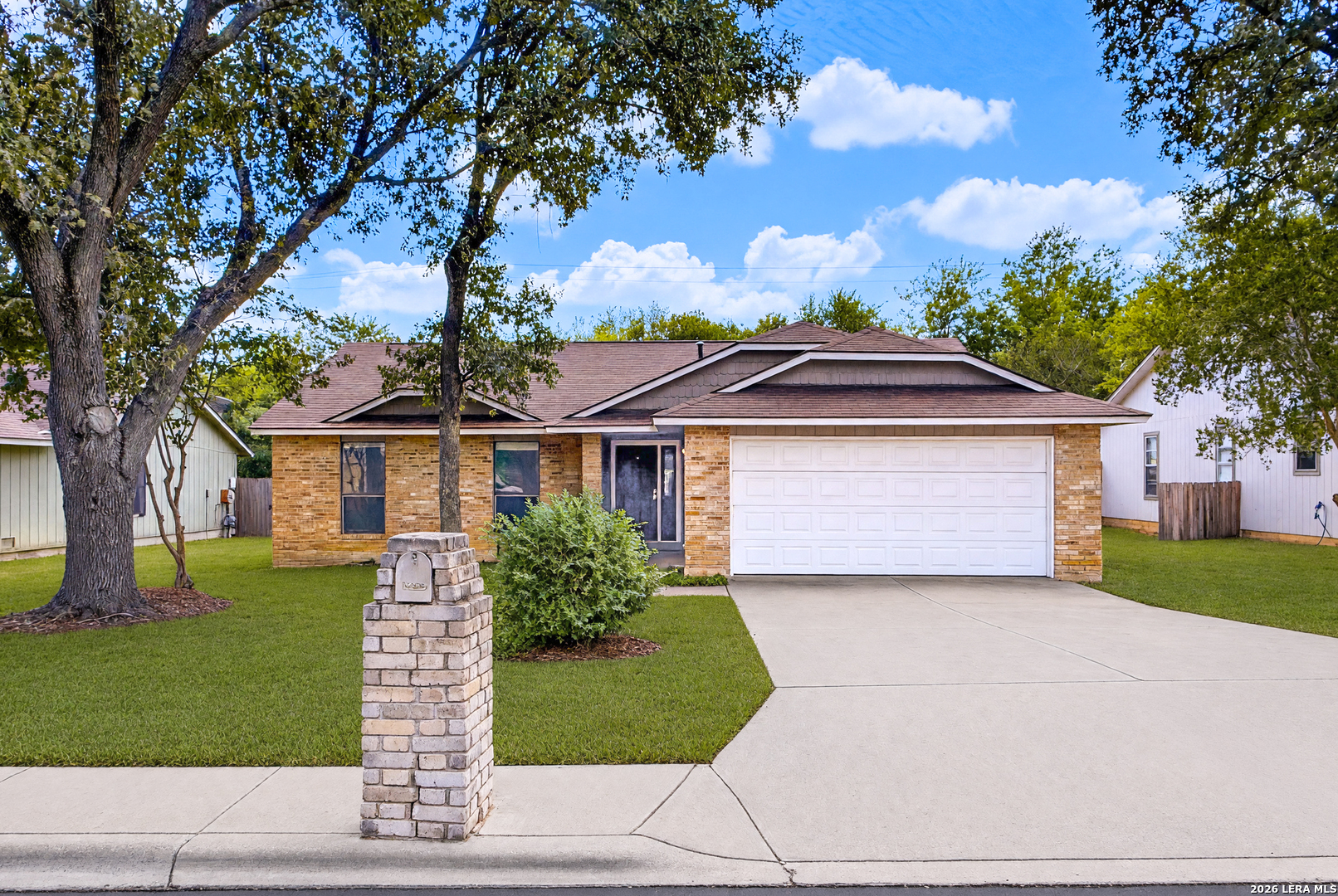 Single-story brick house with a two-car garage, shaded lawn, and brick mailbox pillar.