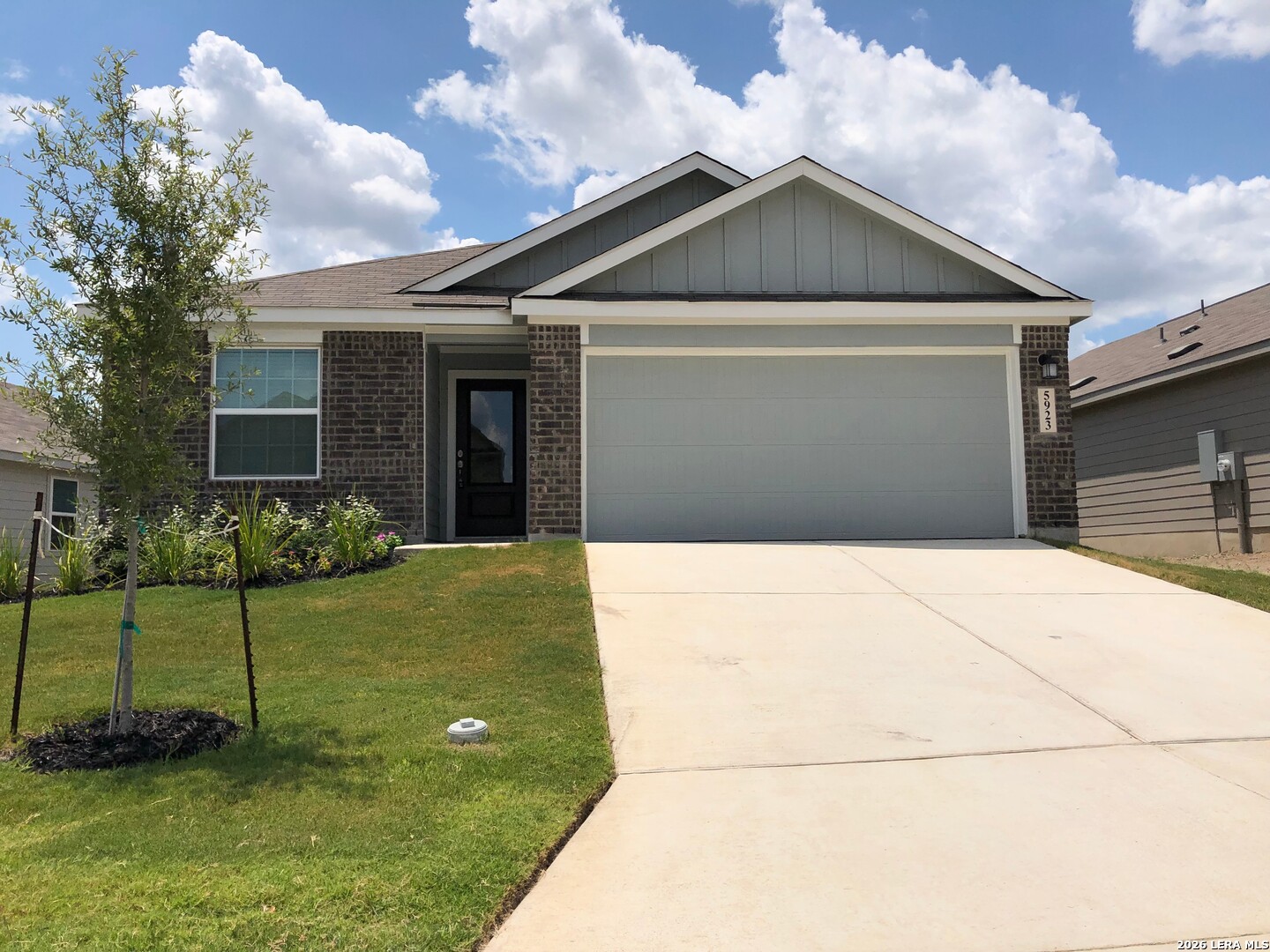 Single-story brick and gray house with a wide garage, concrete driveway, and young tree in the front lawn.
