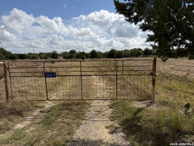 Rural vacant land behind a metal gate marked 1887, with a gravel track and open fields.