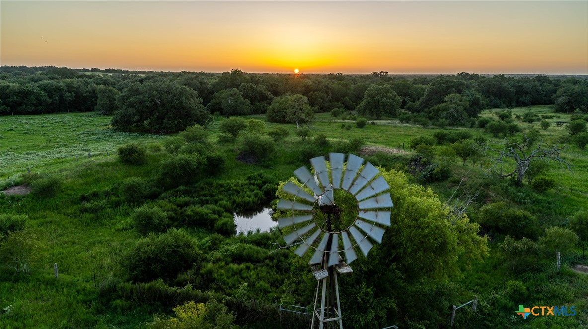 Aerial view of green ranchland with a windmill and pond at sunset.