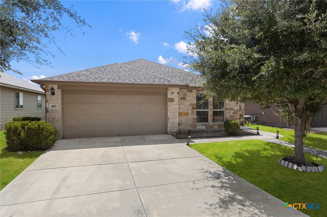 Single-story stone-front home with a wide garage, concrete driveway, green lawn, and mature tree.
