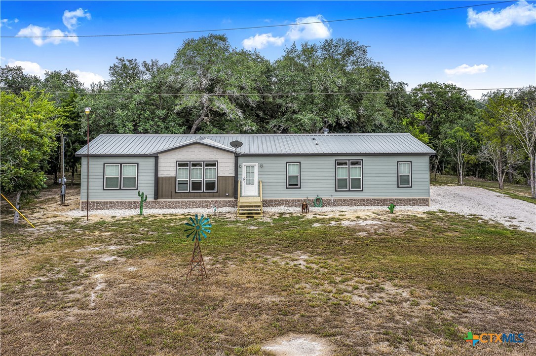Single-story gray home with a metal roof, gravel driveway, and small windmill in the yard.