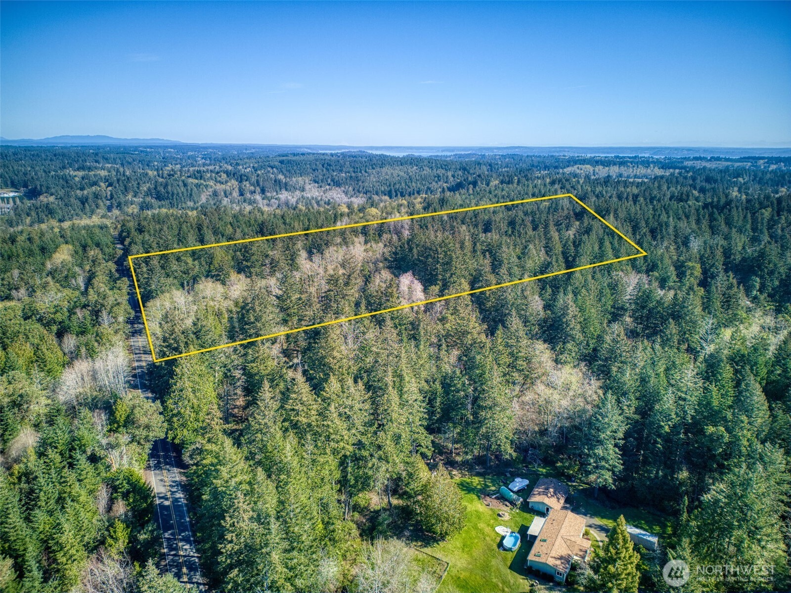 Aerial view of a forested parcel outlined in yellow beside a road, with a neighboring home nearby.