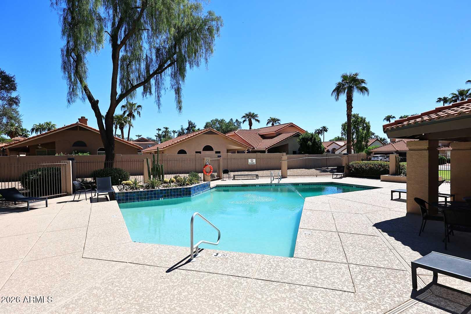 Outdoor community pool with a pale stone deck, lounge chairs, palm trees, and red tile roofs.