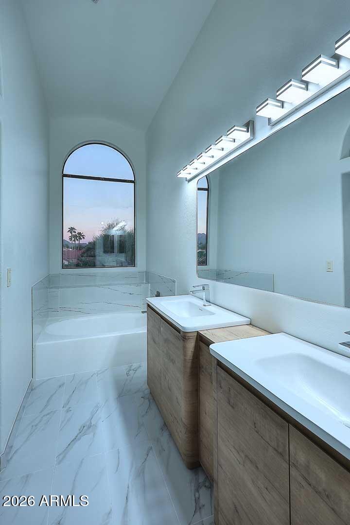 Bathroom with a soaking tub beneath an arched window and a wood double vanity with two sinks.