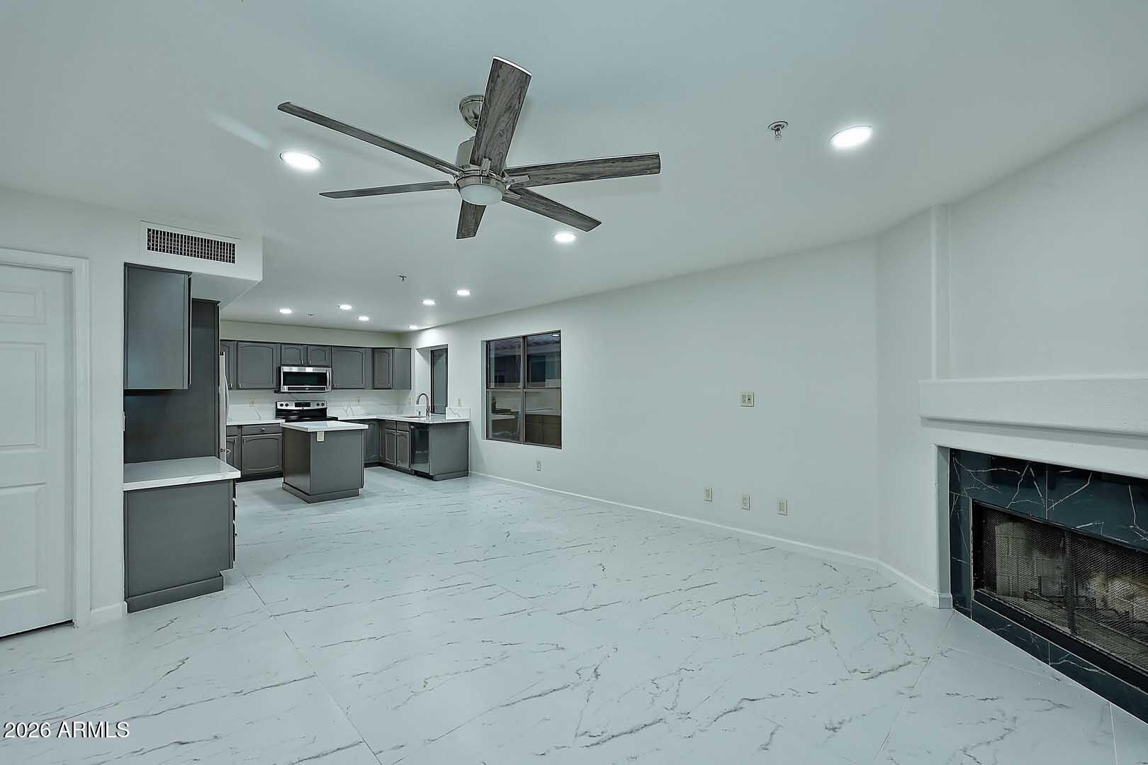 Empty living room and kitchen with gray cabinets, marble-look floors, and a dark stone fireplace.