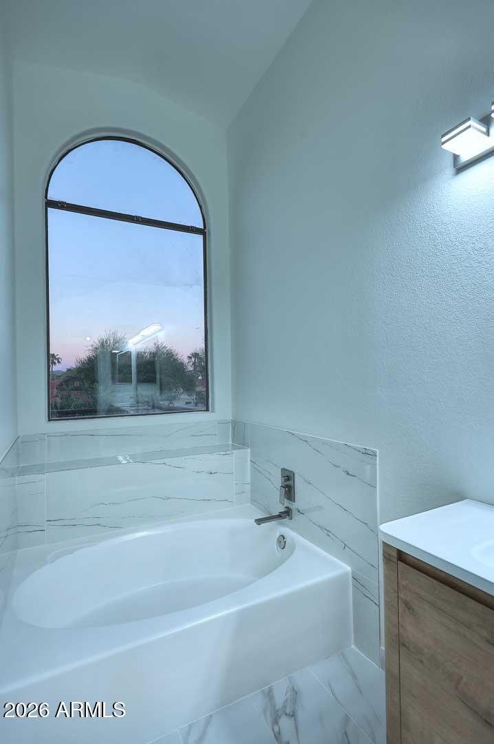 Bathroom with a white soaking tub beneath an arched window and marble-look tile surround.
