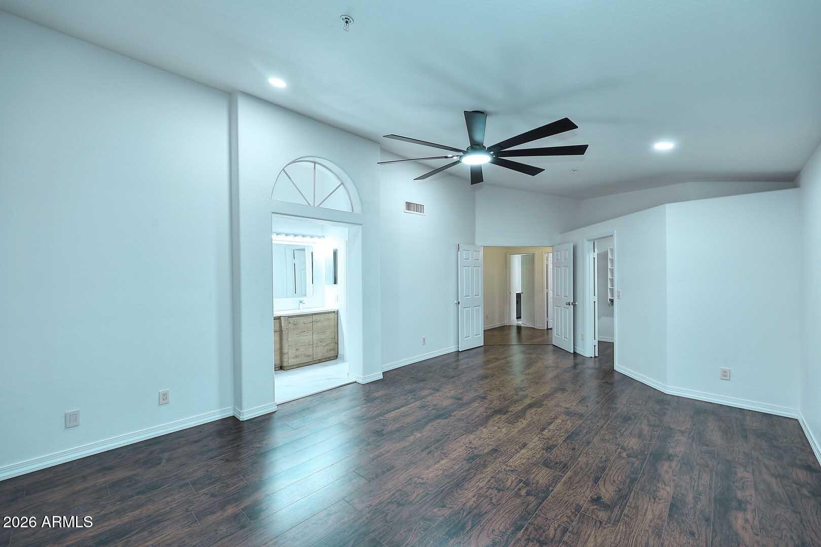 Vacant bedroom with dark wood floors, a large ceiling fan, and an arched window above the bathroom entrance.