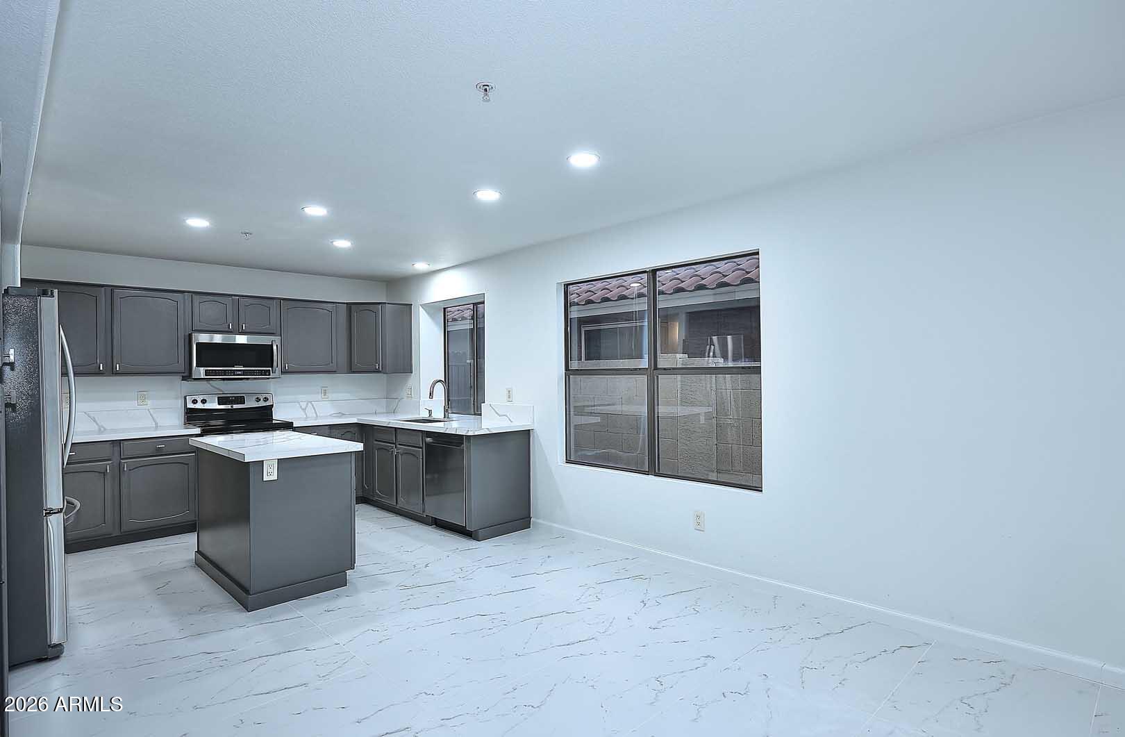 Vacant kitchen with dark gray cabinets, a central island, white veined surfaces, and stainless steel appliances.