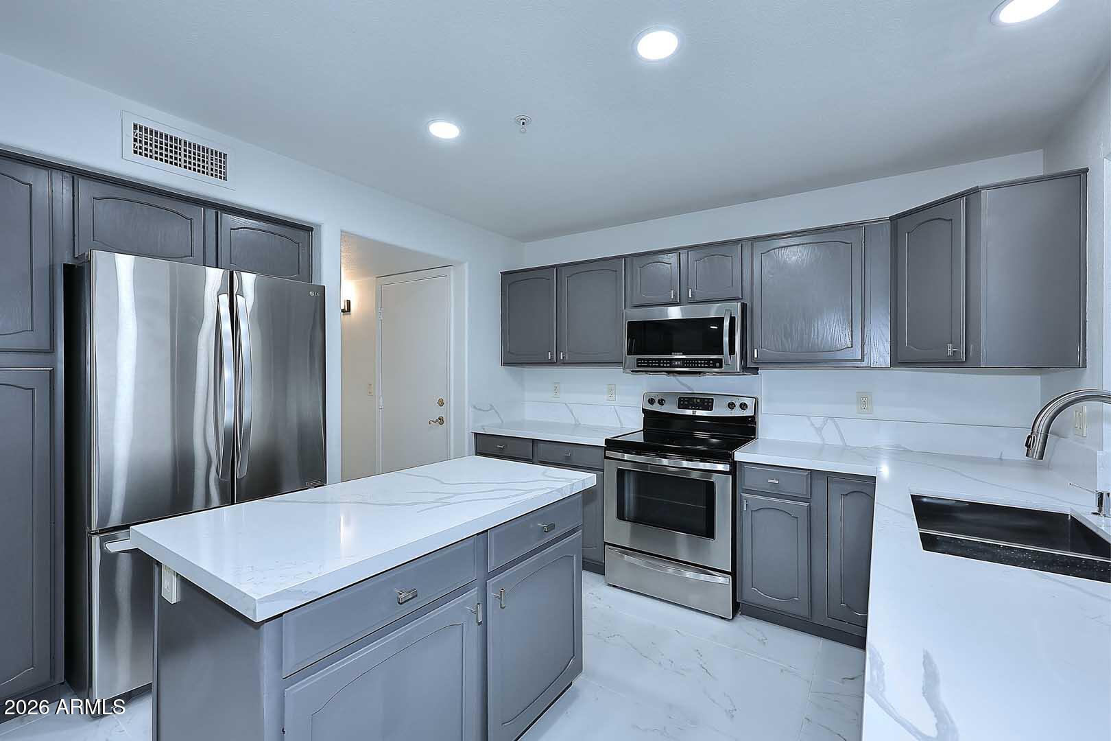 Kitchen with gray cabinets, a large white island, stainless steel appliances, and a black sink.