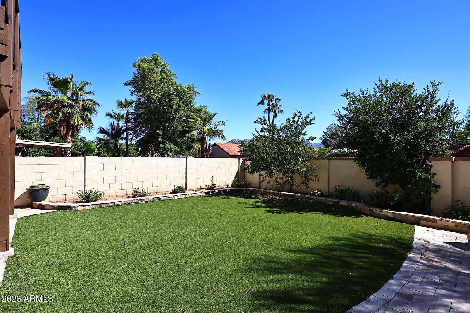 Backyard lawn bordered by a block wall, mature trees, palms, and a paved path.