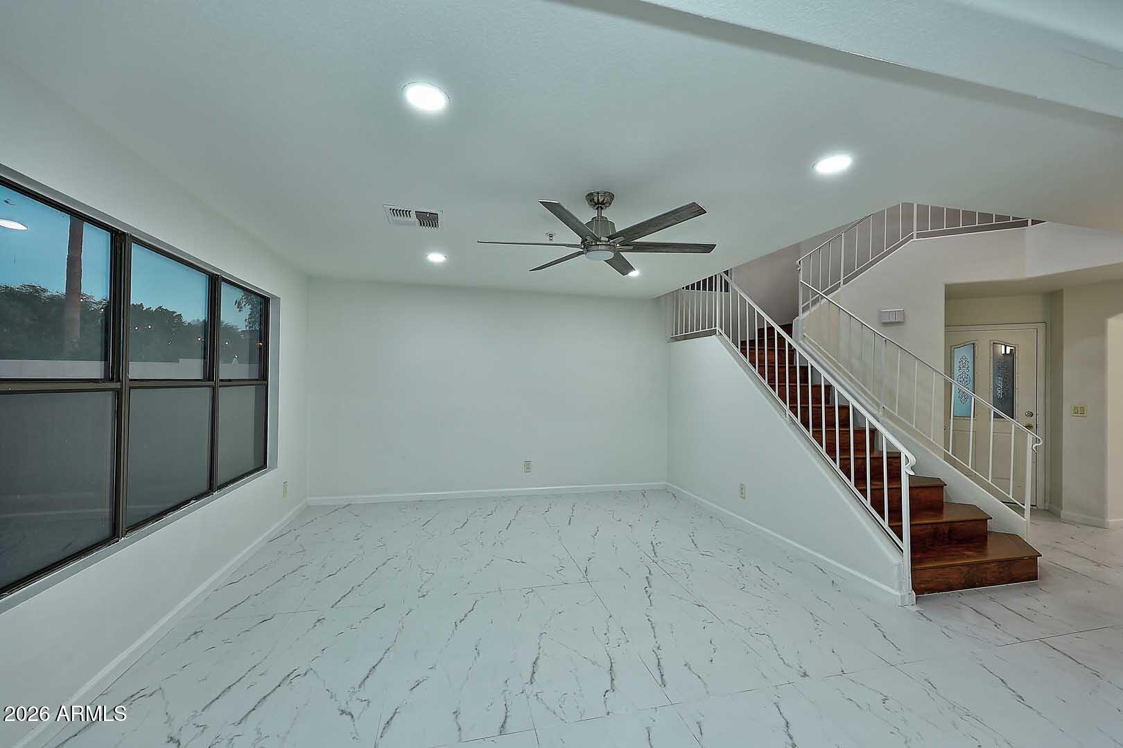 Empty living area with marble-look tile, large windows, and a dark wood staircase.