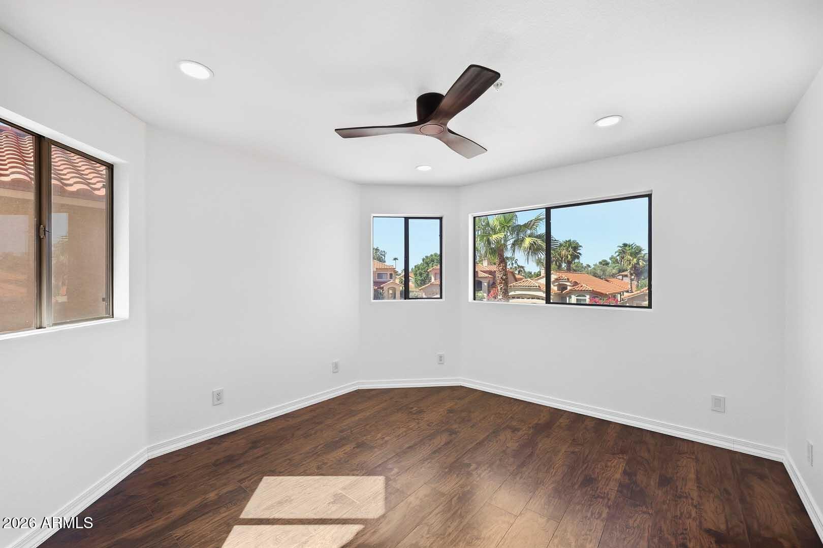Empty room with dark wood floors, a ceiling fan, and windows overlooking palm trees and neighboring homes.