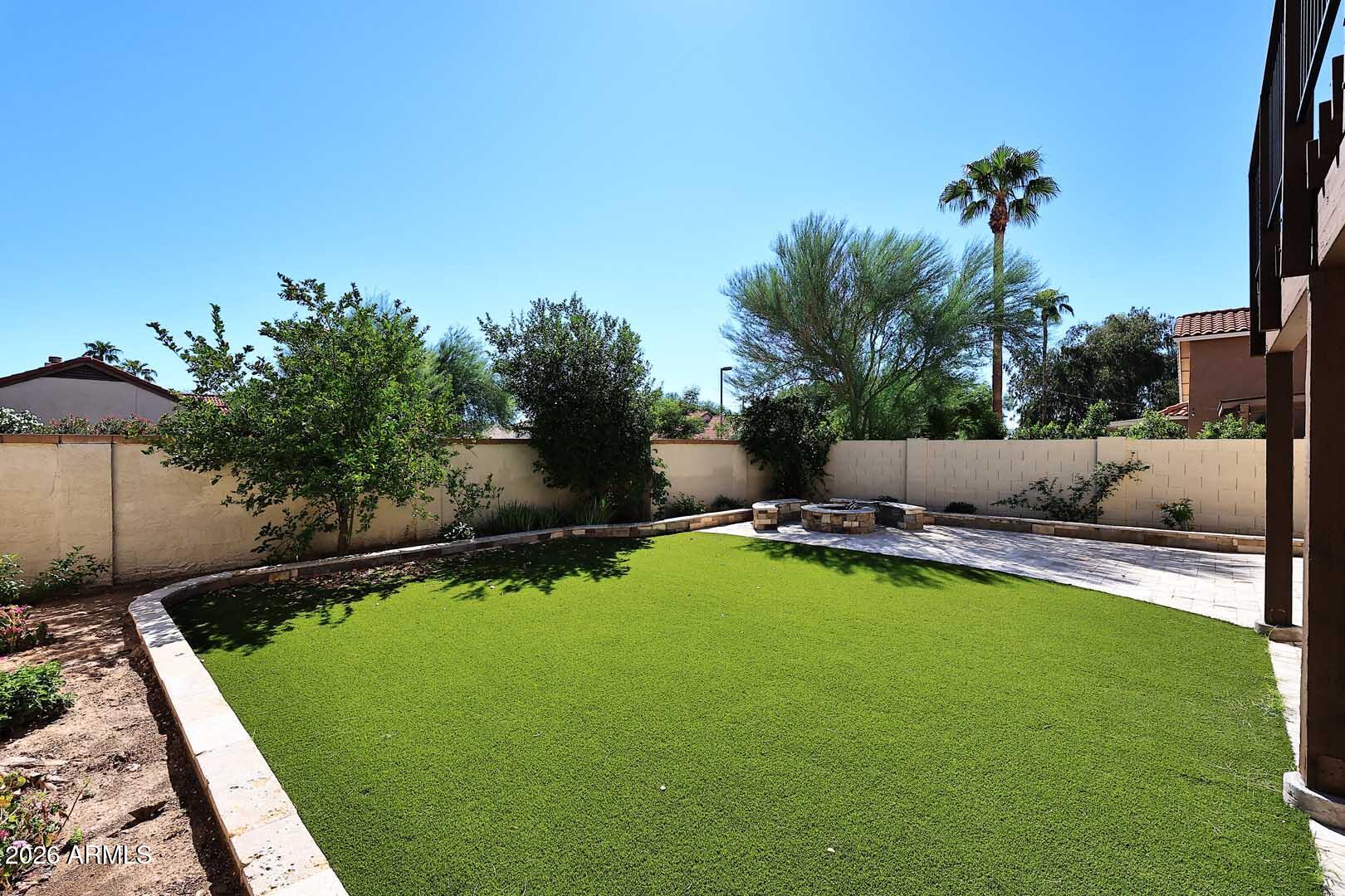 Backyard lawn and paved patio with a circular stone fire pit and mature trees.
