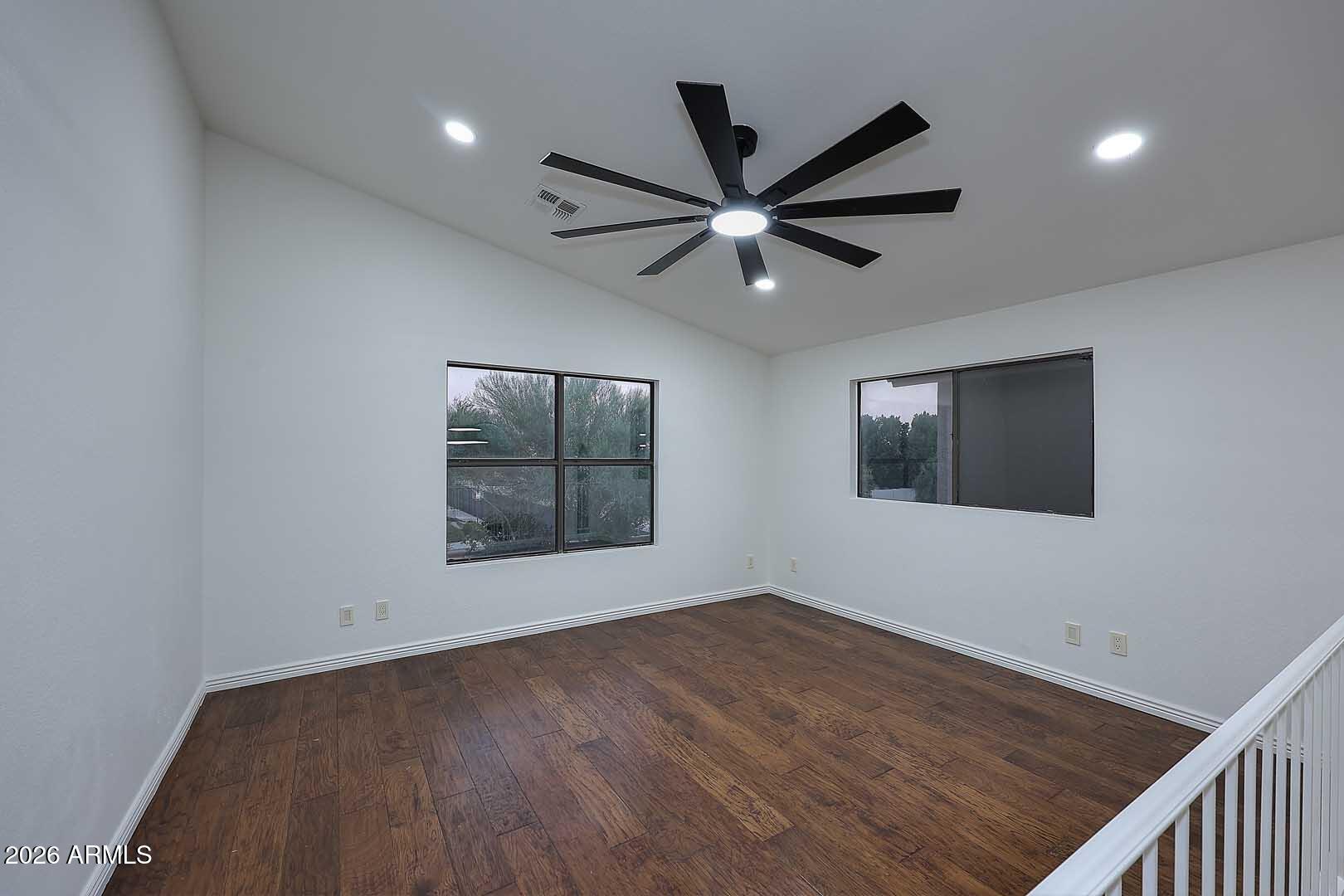 Empty upstairs room with dark wood floors, two windows, and a large black ceiling fan.