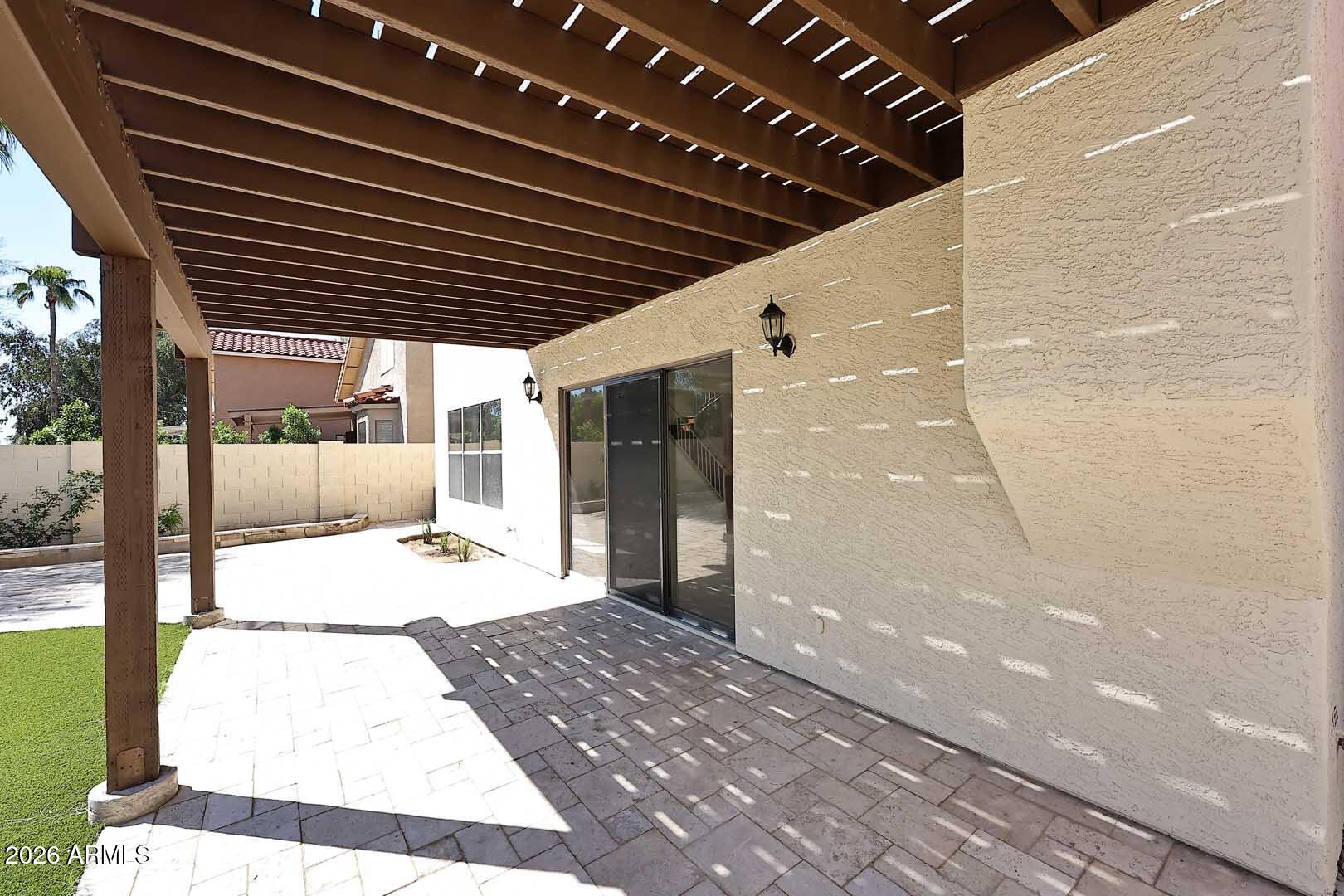 Paved backyard patio beneath a wooden pergola, beside a sliding glass door and artificial turf.