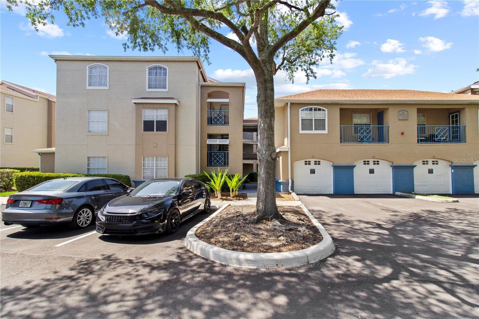 Beige condominium buildings with balconies and garage doors, shaded by a large tree beside the parking lot.