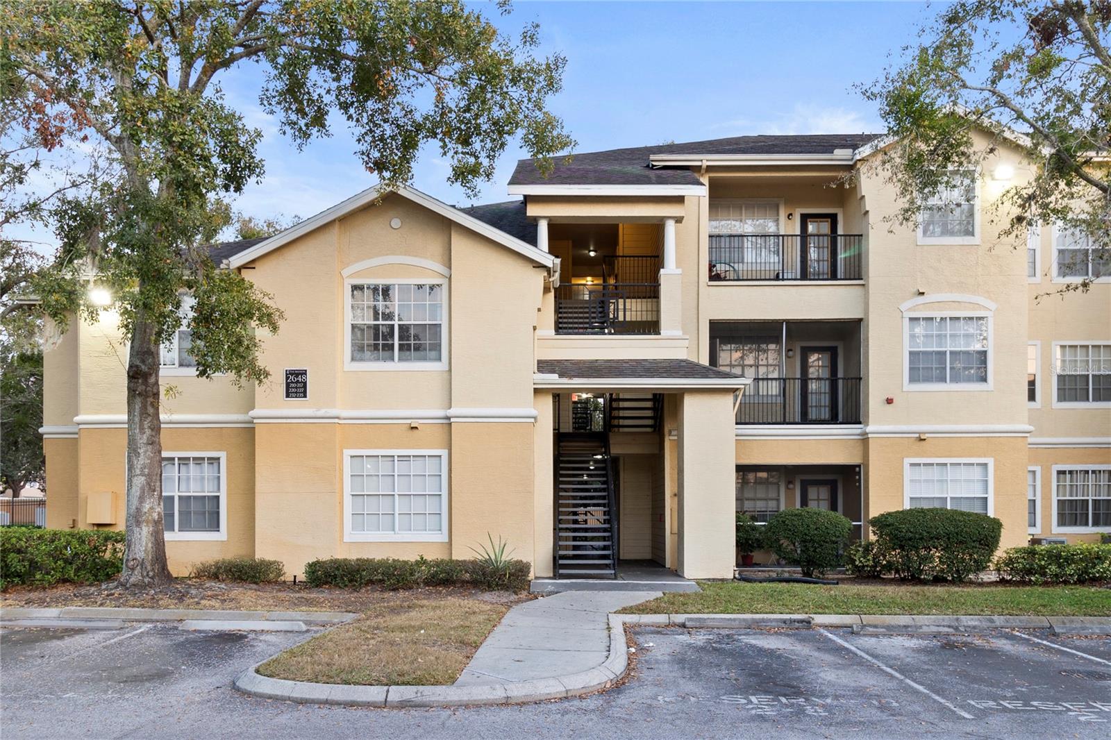 Three-story beige condominium building with exterior stairways, balconies, and a landscaped entrance.