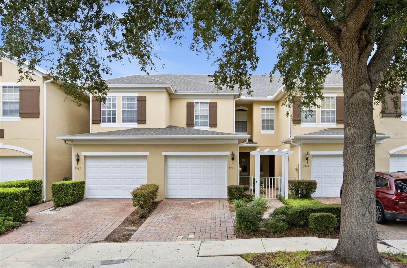Two-story tan condo with brown shutters, white garage doors, and a brick-paver driveway beneath a large tree.