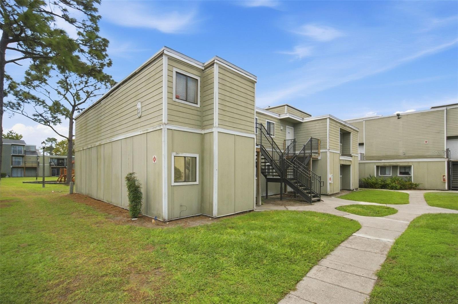 Beige two-story condo building with exterior staircases beside a curved walkway and lawn.