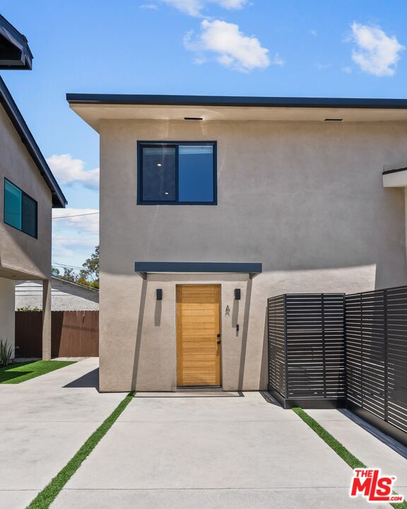 Modern stucco home with a wood entry door, concrete driveway, and black slatted privacy fence.