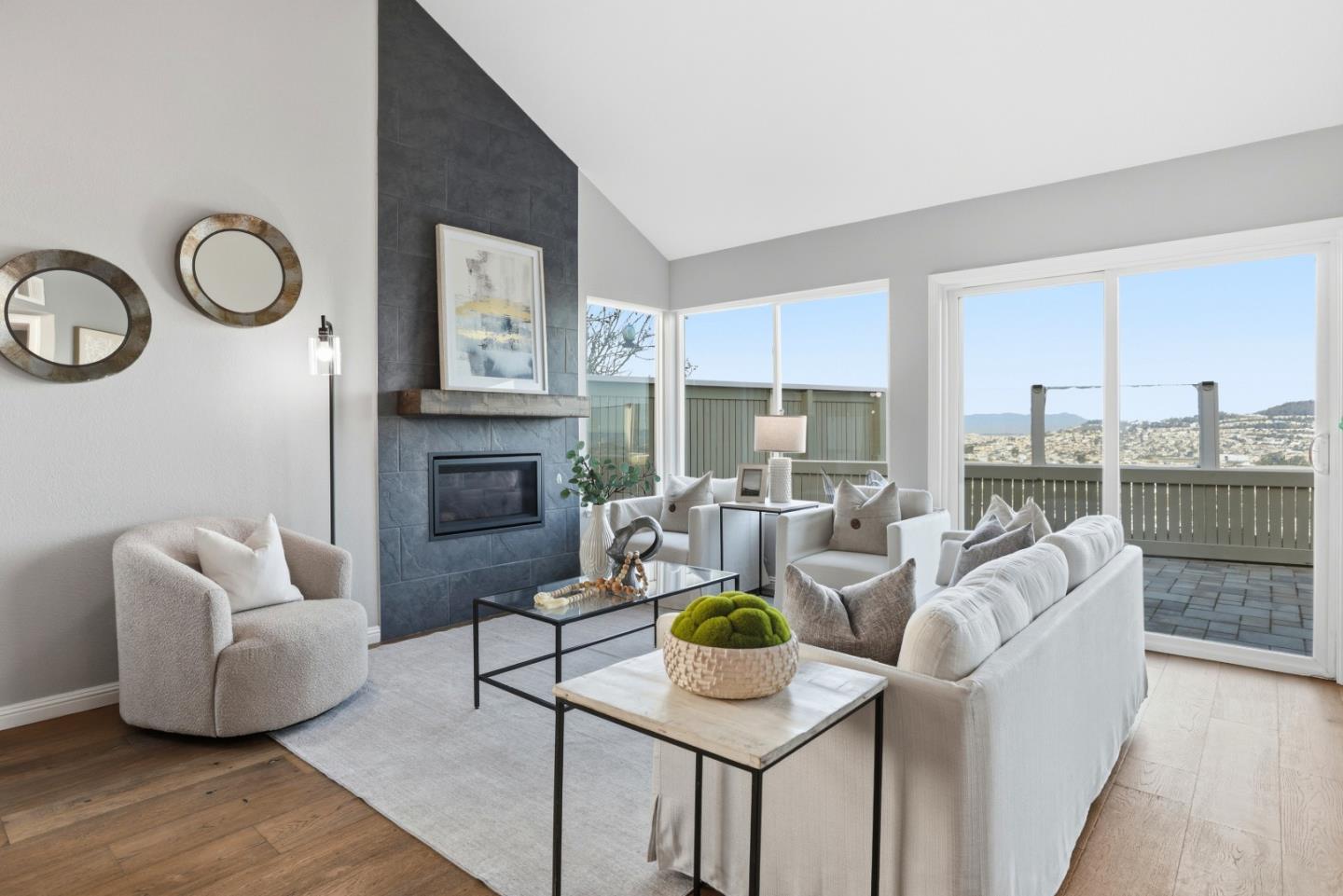 Living room with a dark tile fireplace, white seating, and windows overlooking hillside homes.