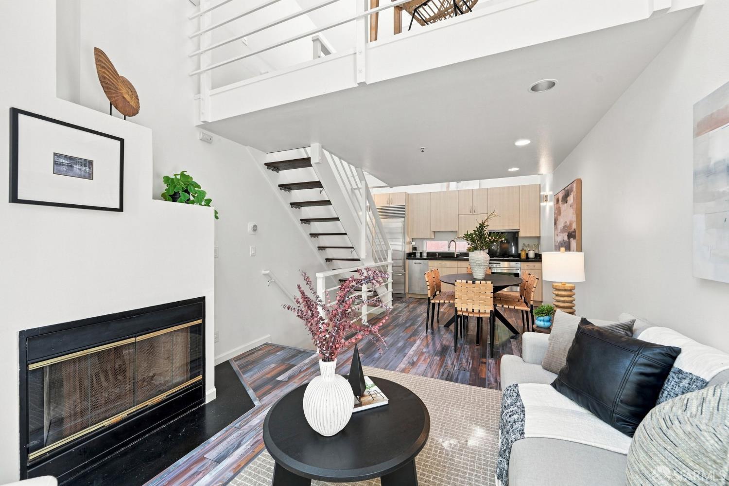 Open-plan living room with a black fireplace, multicolored wood floors, dining area, and staircase to a mezzanine.