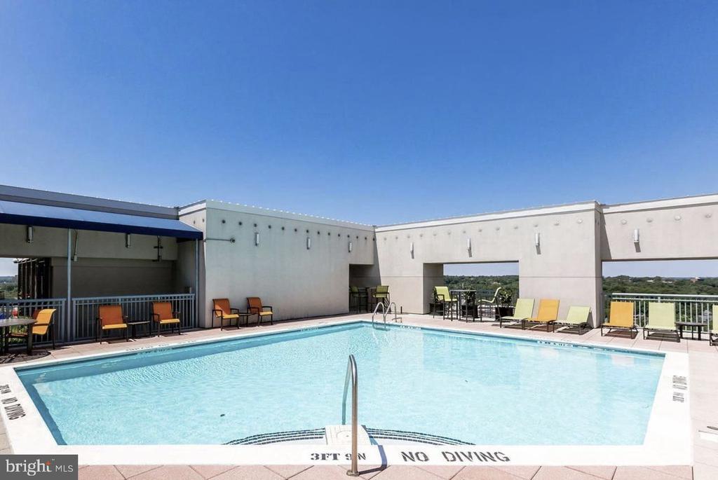 Rooftop pool with lounge chairs, tables, and distant tree views.