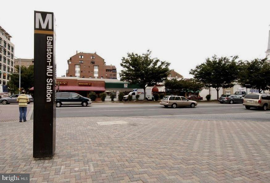 Ballston-MU Station sign beside an urban street with shops and passing cars.