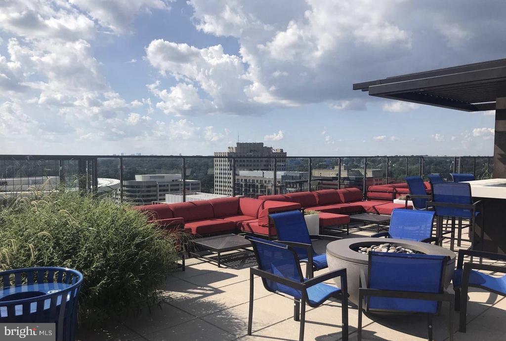 Rooftop terrace with red sectional seating, blue chairs, and a fire pit overlooking nearby buildings.