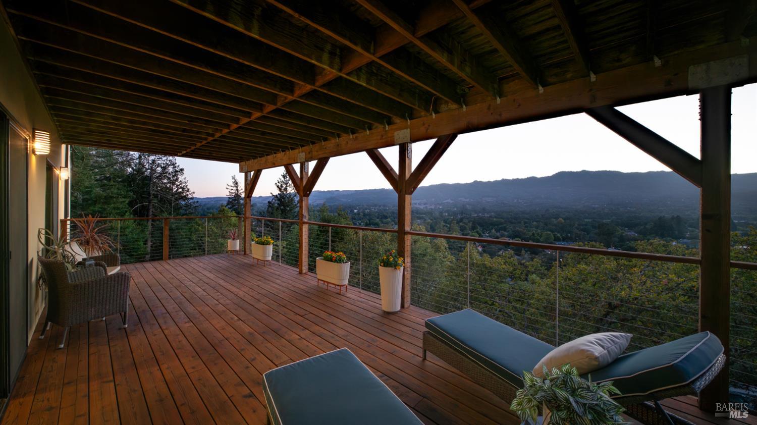 Covered wooden deck with loungers and potted plants overlooking a wooded valley and distant mountains.