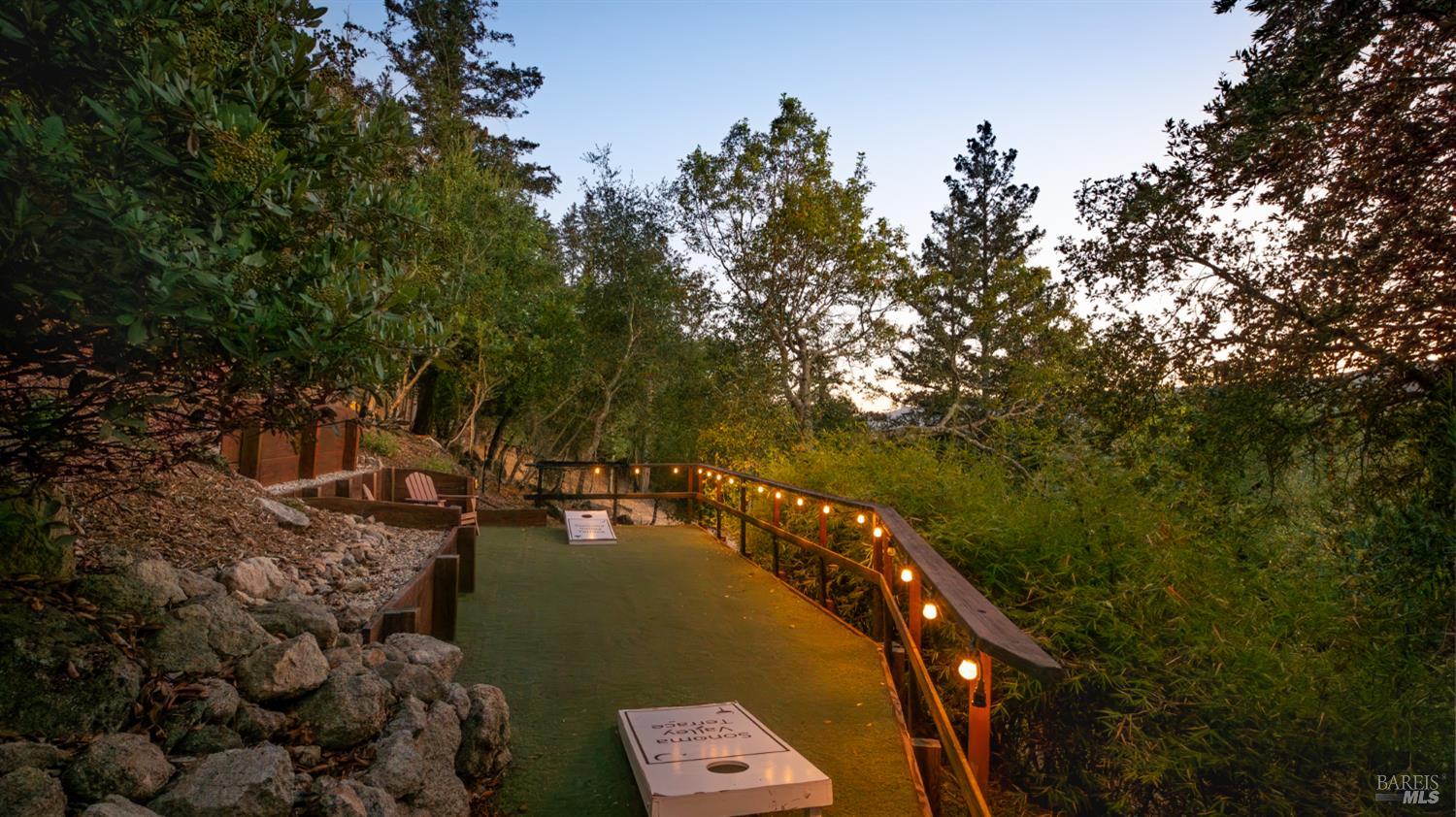 Wooded cornhole court with seating and string lights along the railing at dusk.