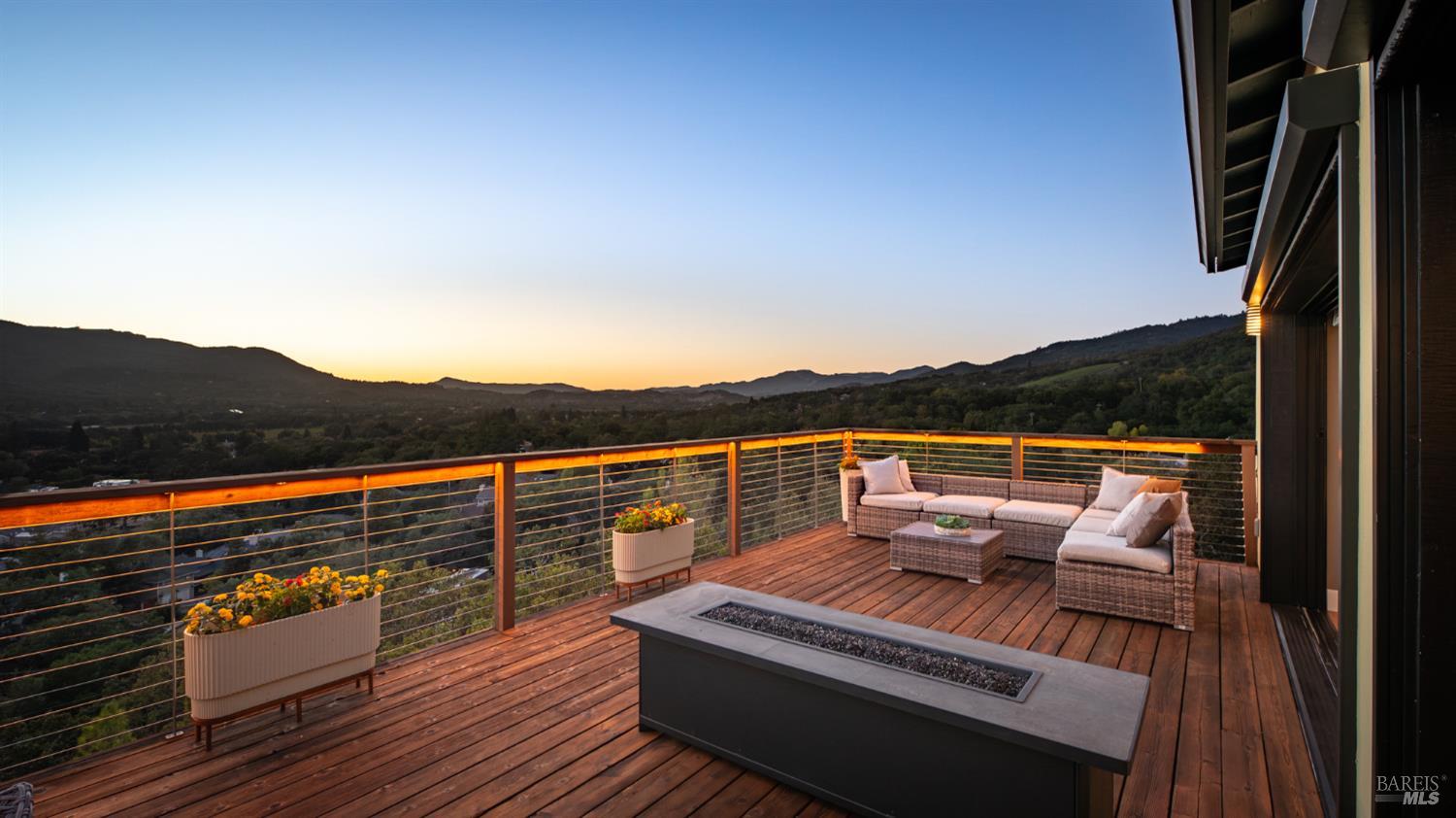 Furnished wood deck with a built-in fire feature overlooking a wooded valley at sunset.