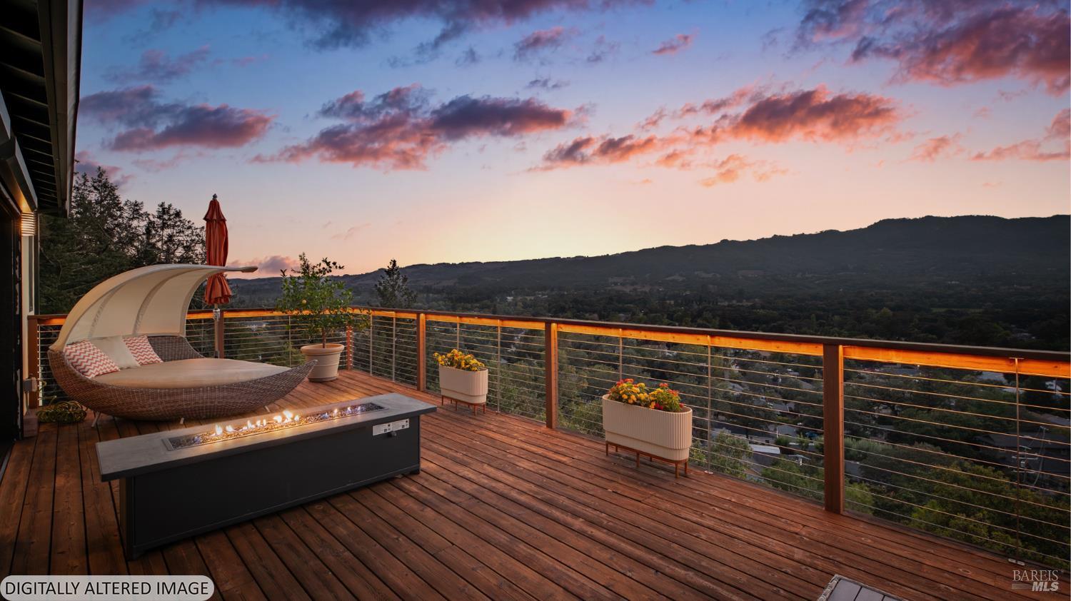 Furnished wooden deck with a fire table overlooks wooded hills beneath a pink and blue dusk sky.