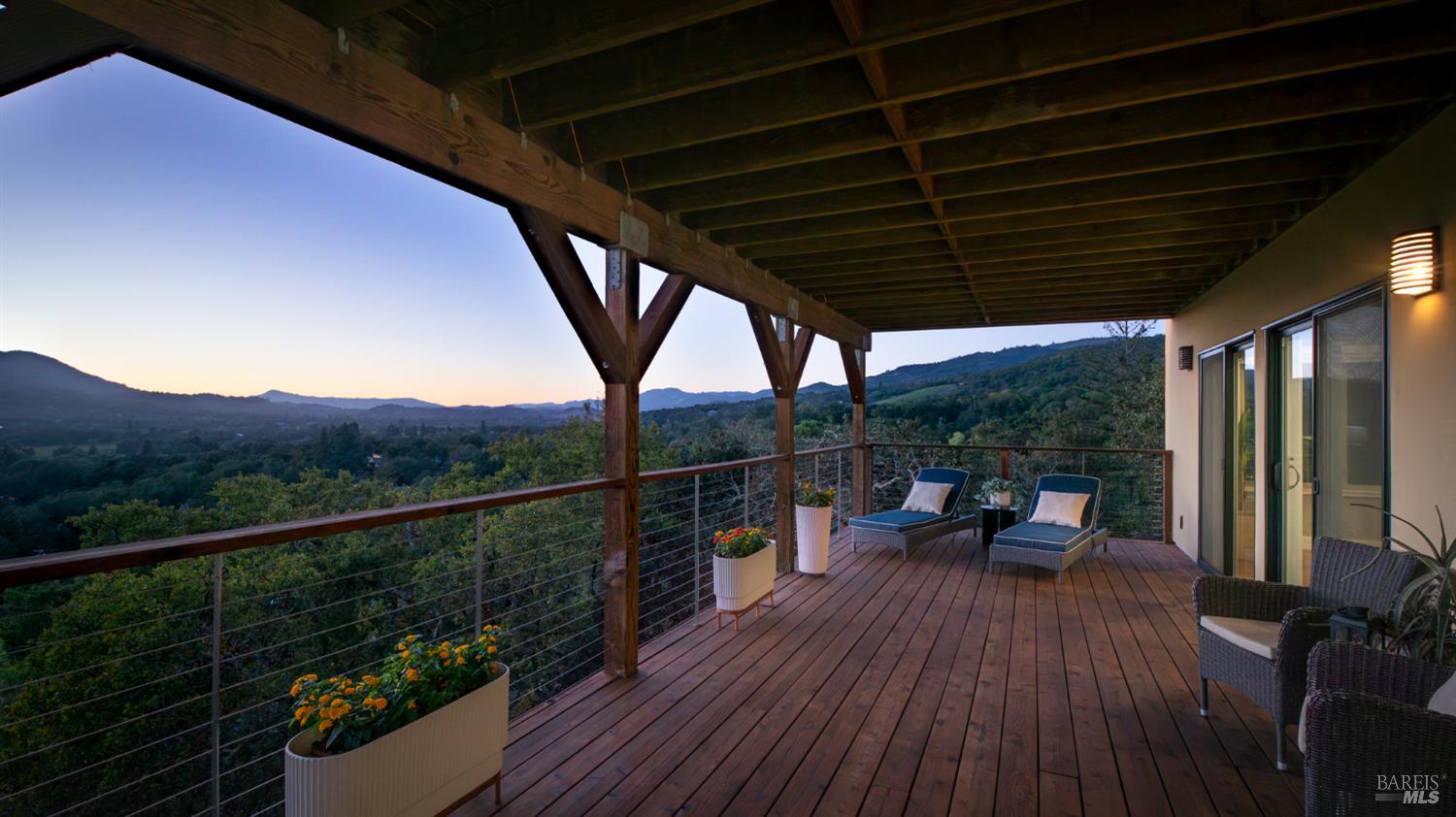 Covered wooden deck with lounge chairs overlooking a wooded valley and distant hills at dusk.
